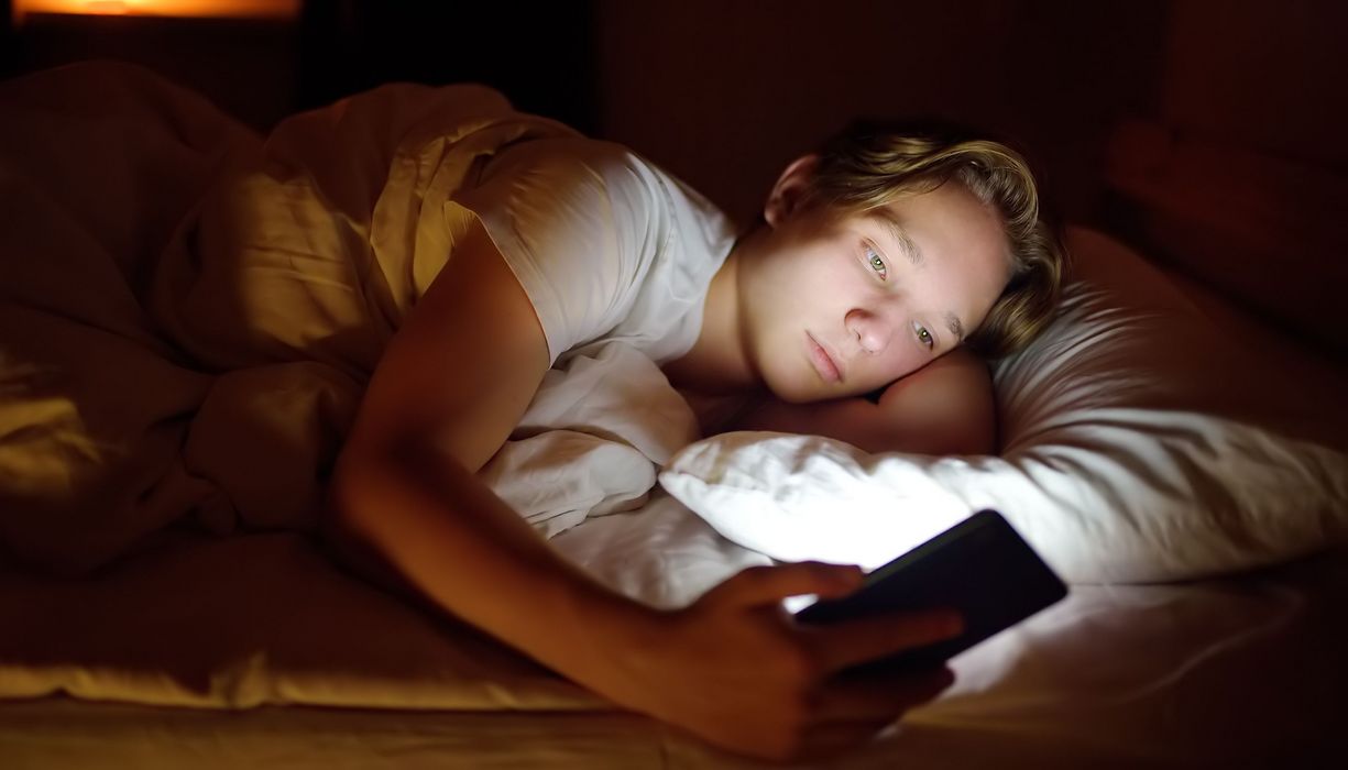 Jugendlicher, liegt im Bett und schaut auf sein Smartphone.