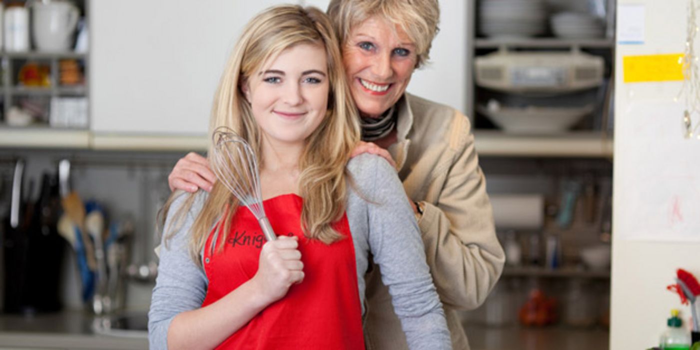 Küchenszene: Teenagertochter im Vordergrund, blonde Haare, rote Schürze, Schneebesen in der rechten Hand, dahinter sie an den Schultern haltend Mutter, beide in die Kamera lächelnd