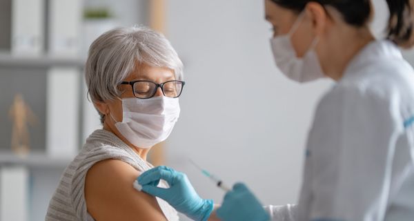 Ältere Frau wird von einer jungen Ärztin geimpft, beide tragen eine Maske.