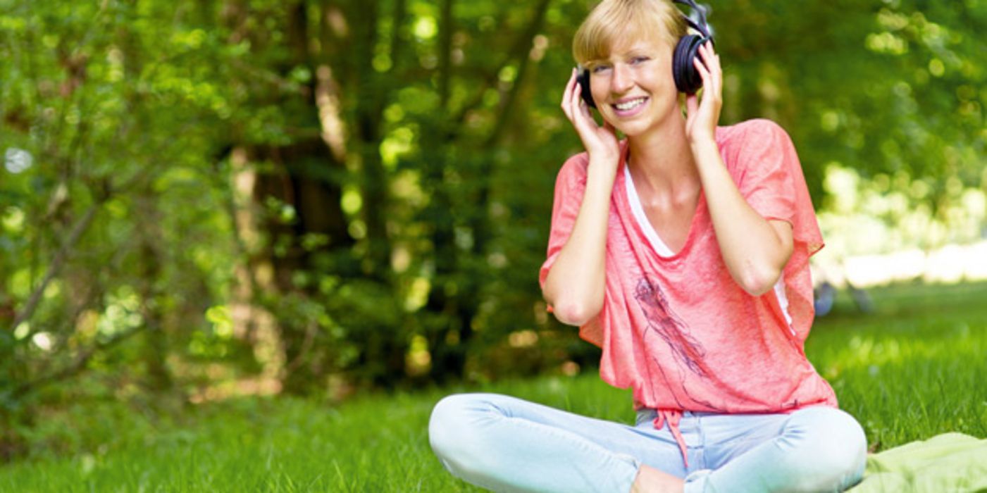 Junge Frau sitzt auf Decke im Park und hört Musik über Kopfhörer, lacht in die Kamera