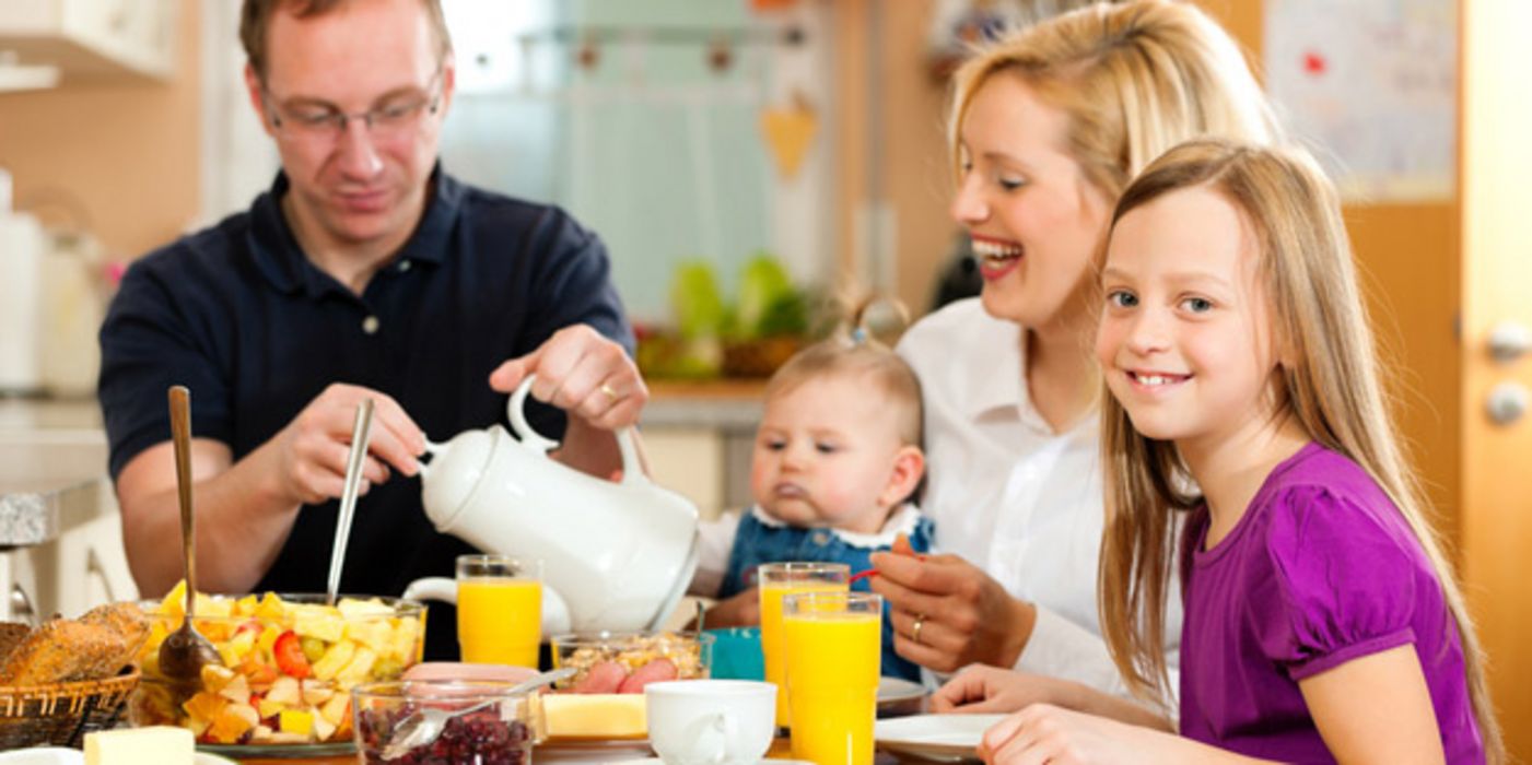 Mutter, Vater, Tochter (ca. 8 Jahre alt) und Baby beim vergnügten Frühstück, blonde Tochter im Vordergrund lächelt in die Kamera