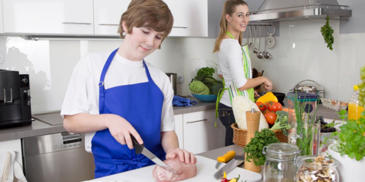 Geschwister im Teenager-Alter stehen in der Küche, der kleinere Bruder schneidet Fleisch, seine ältere Schwester wäscht Gemüse ab