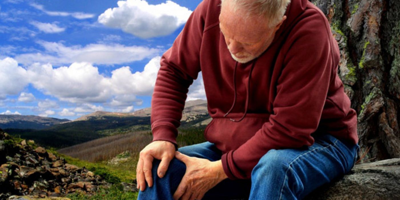 Senior in den Bergen auf Fels sitzend und sich ans rechte Knie greifend