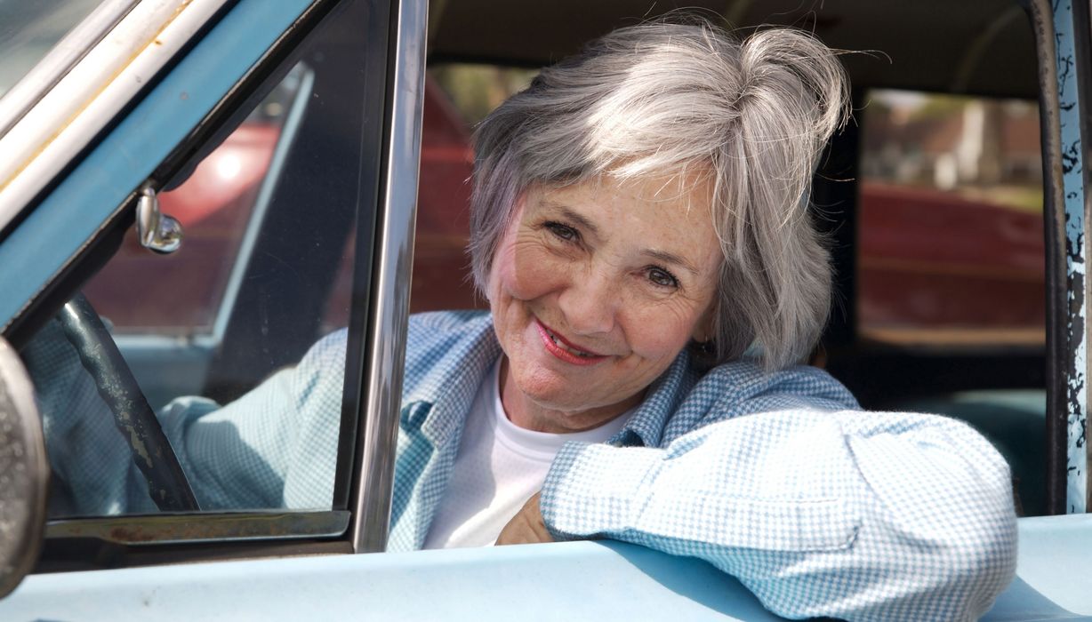 Ältere Frau, lächelt aus dem Autofenster heraus.