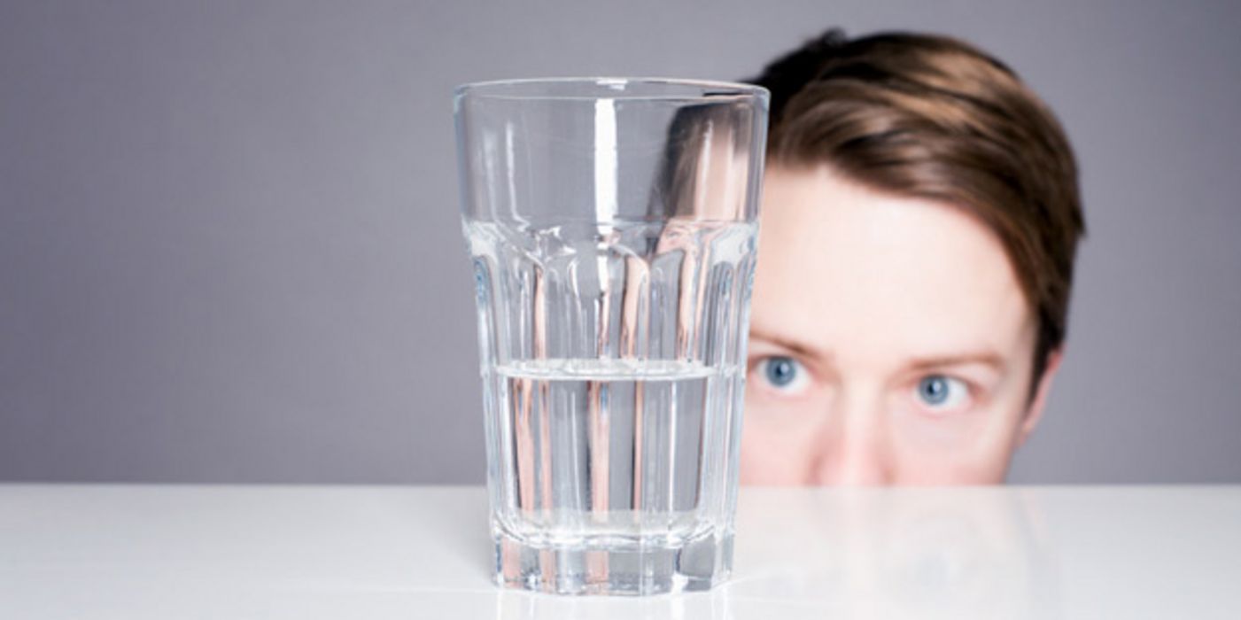 Frau lugt über eine Tischkante auf ein halb-volles Wasserglas auf dem Tisch