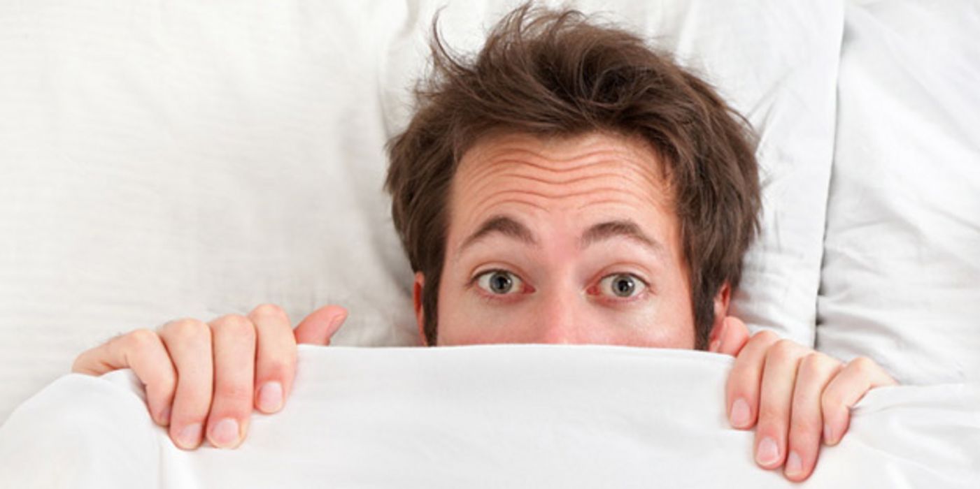 Mann liegt unter einer Bettdecke; nur der obere Teil des Kopfes lugt hervor; die Augen blicken angsterfüllt