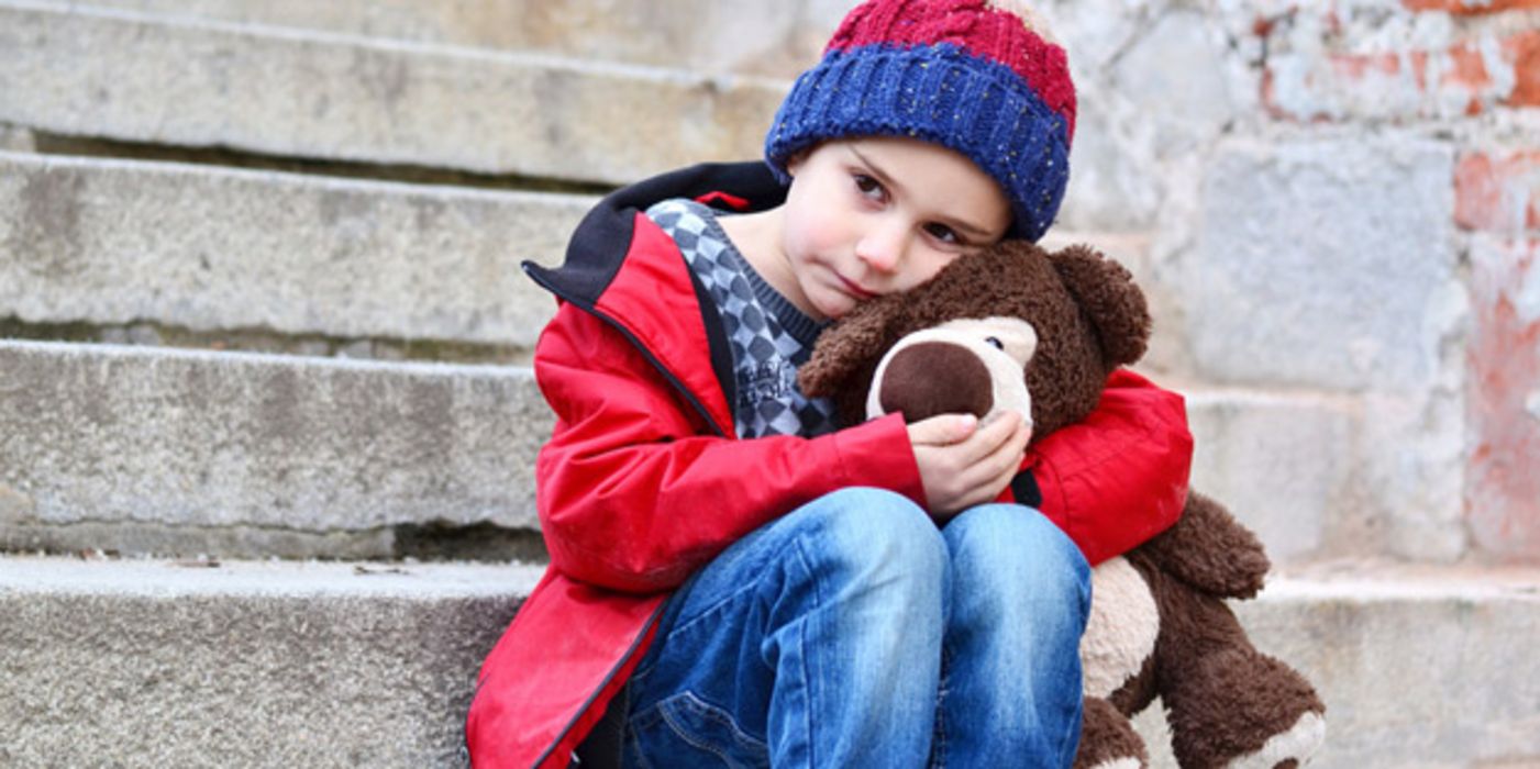 Kindergartenkind, ca. 5 Jahre alt, Mütze, Anorak, Jeans, einen Teddy im Arm, sitzt traurig auf einer grauen Steintreppe