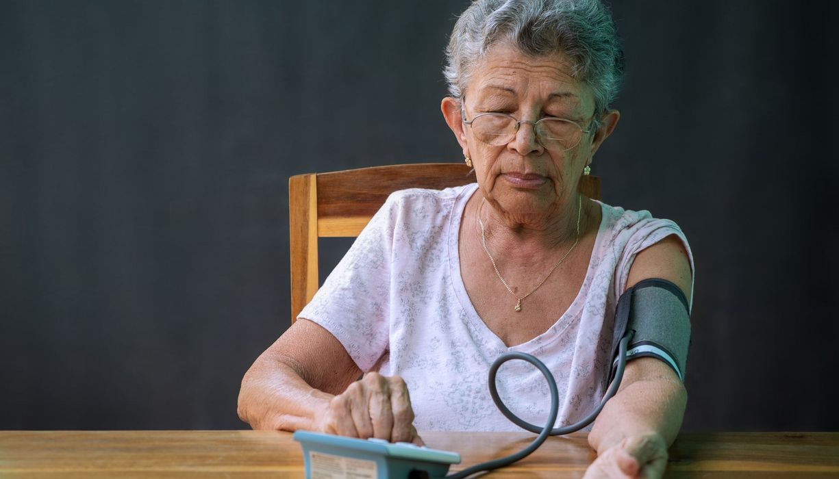 Ältere Frau, ca. 80 Jahre alt, misst mit einem Blutdruckgerät am Oberarm ihren Blutdruck.
