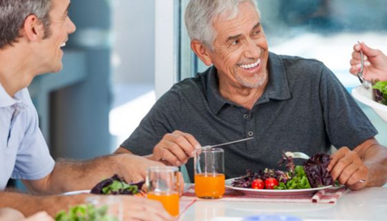 Zwei attraktive Männer über 60, grauhaarig, lachend, am Tisch mit Salatteller und Saft
