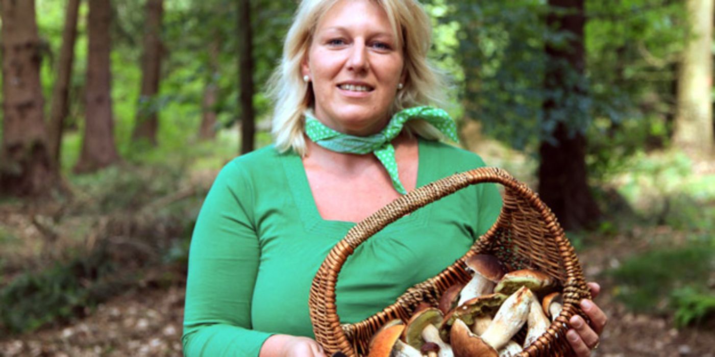 Frontalfoto, Frau in den 40ern, blond, türkises Langarmshirt, türkises Nickituch, im Wald, Korb mit Pilzen in den Händen