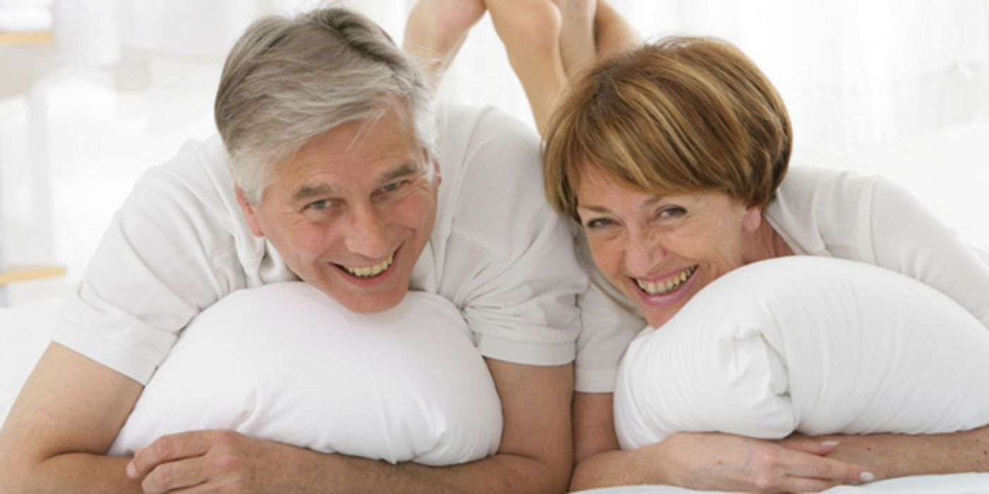 Vitales Seniorenpaar liegt bäuchlings im Bett und lacht in die Kamera