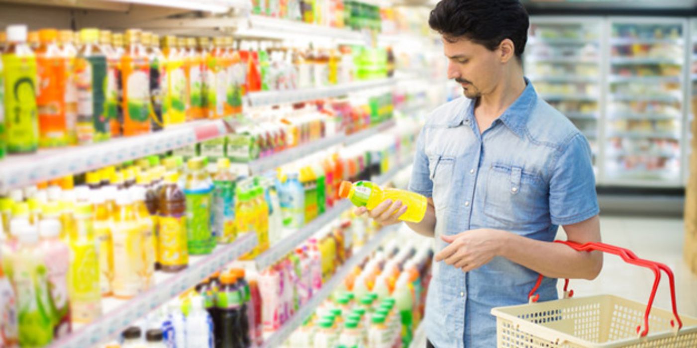Mann um die 30 im Sommerhemd mit Einkaufskorb am Kühlregal, schaut auf Etikett einer Flasche mit Saft oder Öl