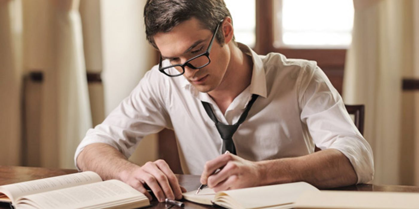 Junger Mann mit Brille, Hemd und Krawatte sitzt an einem Tisch und schreibt mit der linken Hand einen Text