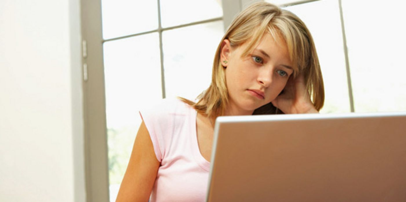 Junge blonde Frau, ca. 20, am Tisch sitzend, schaut skeptisch in Laptop, Kopf aufgestützt