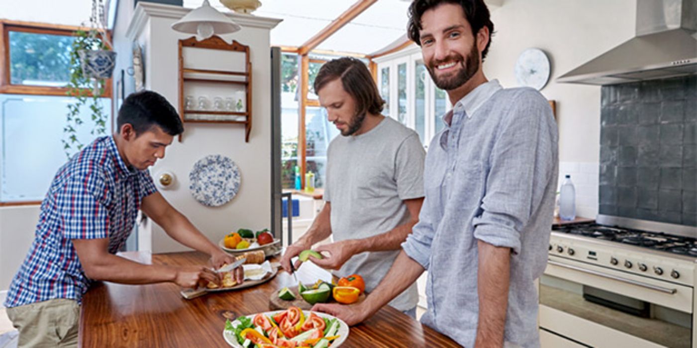 Drei Männer Ende 20 in Freizeitkleidung ín Küche beim Zubereiten einer vegetarischen Mahlzeit, bärtiger Mann im Vordergrund lacht in Kamera