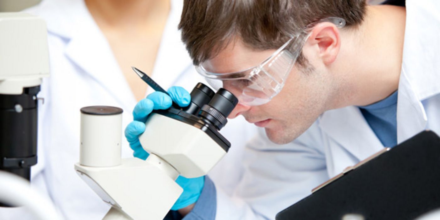 Junger Mann mit weißem Kittel und Schutzbrille schaut durch ein Mikroskop.