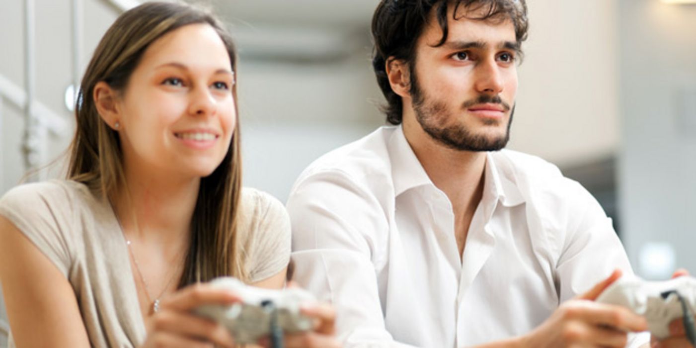 Junge Frau und junger Mann sitzen nebeneinander und halten Spielkonsolen in der Hand