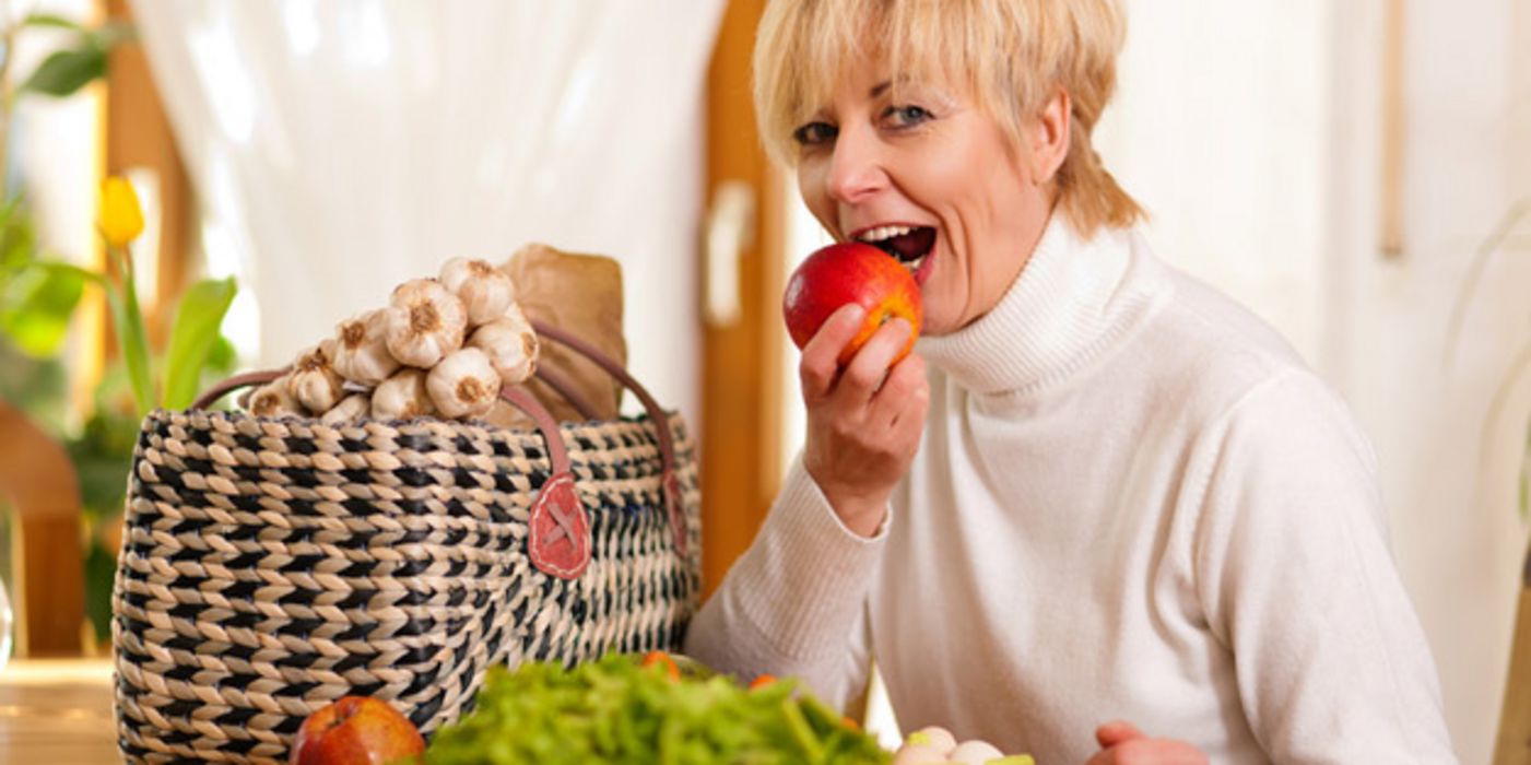 Blonde Frau um die 50 beißt in einen Apfel, auf dem Tisch grüner Salat, Apfel, Möhre und ein Korb, aus dem Knoblauchstränge herausschauen