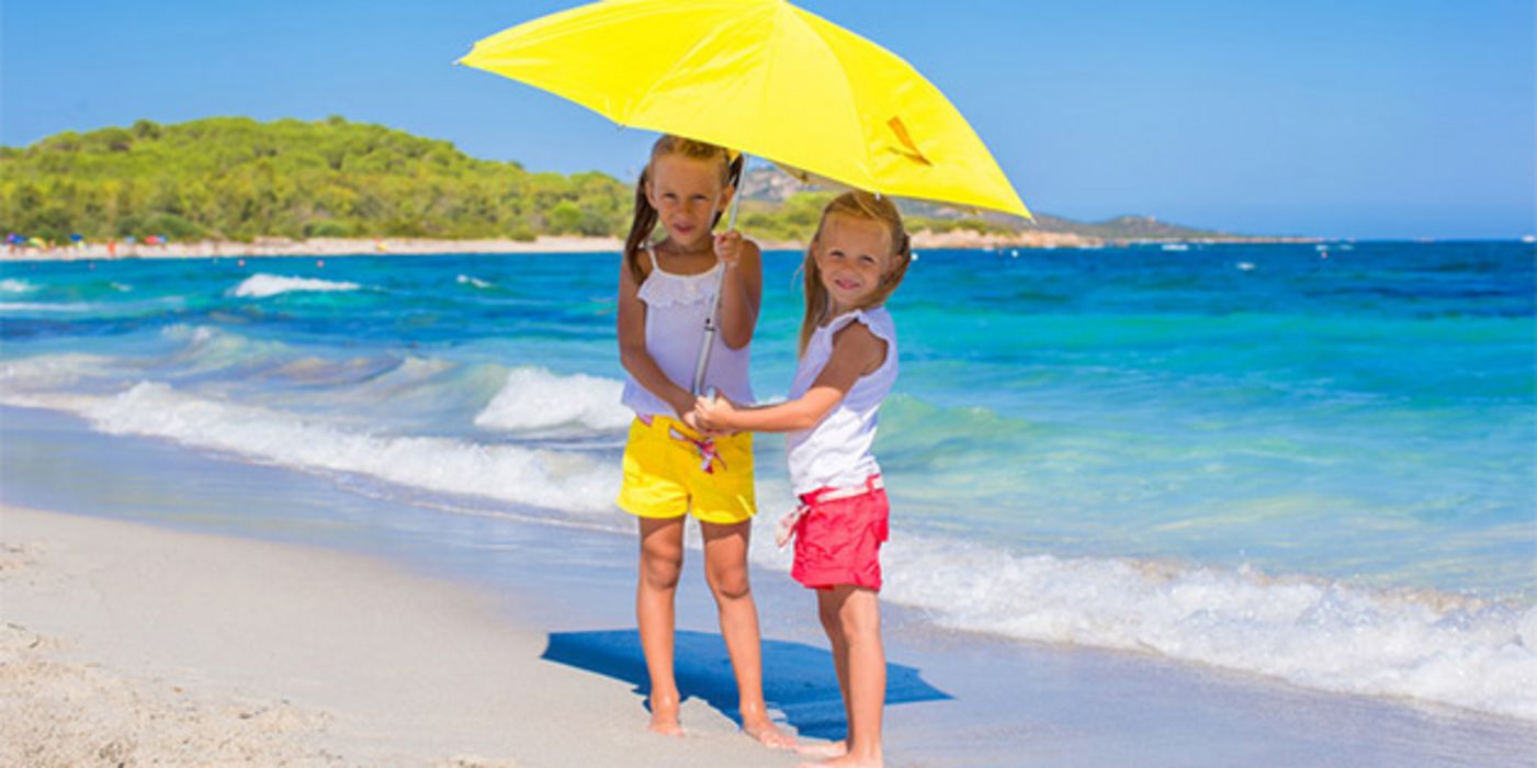 2 blonde Mädchen (ca. 9 und 6 Jahre), weiße Shirts, Shorts gelb bzw. blau, unter großem gelbem Sonnenschirm an weißem Strand am blauen Meer