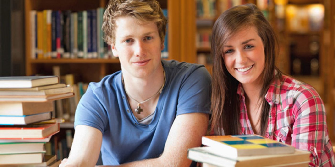 Student und Studentin in einer Bibliothek, umrundet von Bücherstapeln beim Lernen