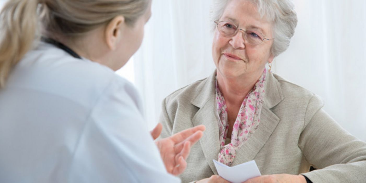 Ältere weißhaarige Dame freundlich lächelnd mit Rezept in der Hand im Beratungsespräch bei Ärztin (rechter Bildrand von hinten seitlich zu sehen)