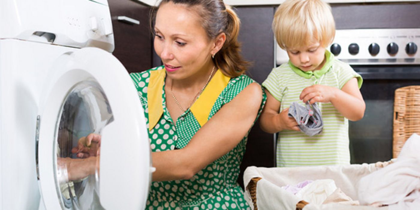 Frau in den 30ern beim Ausräumen der Wäschemaschine im Waschkeller, an ihrer Seite ein Kind, ca. 3 Jahre, mit Wäschestück in den Händen