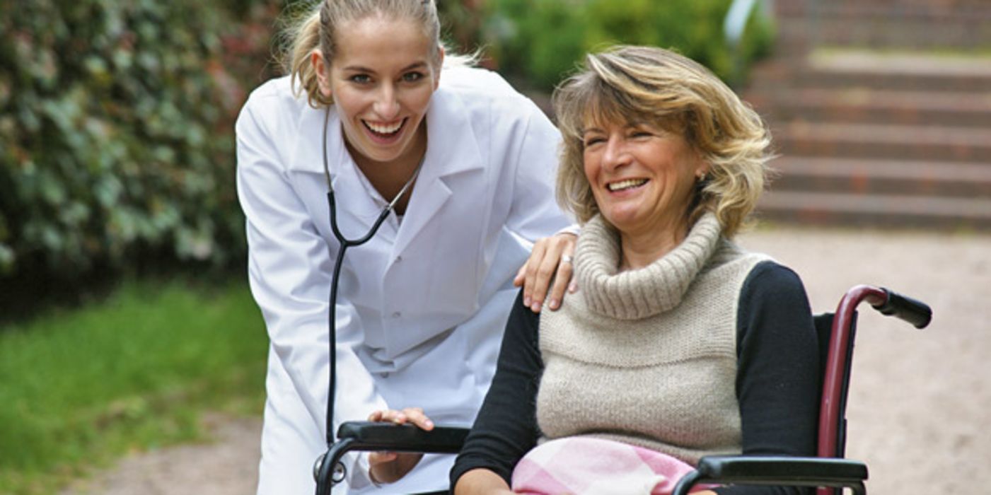 Lächelnde Frau im Rollstuhl im Freien, neben ihr eine junge Ärztin