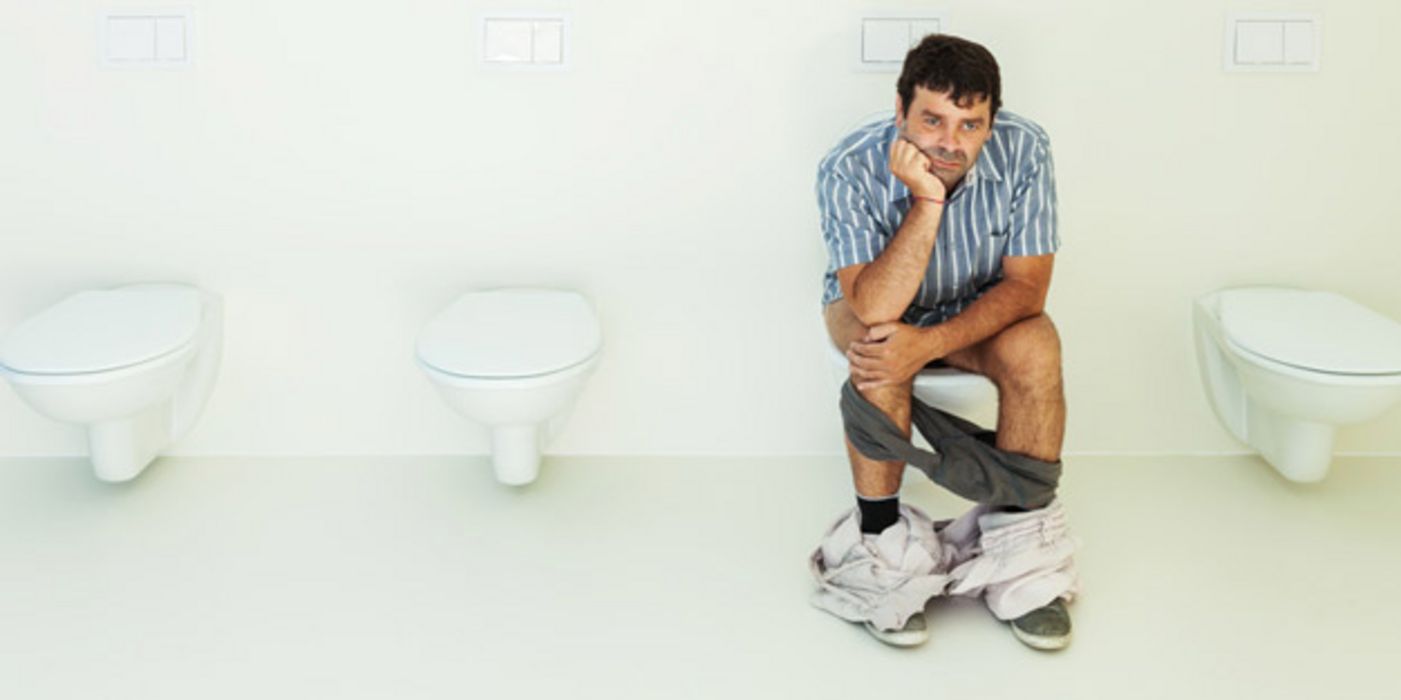 Mann, ca. Anfang/Mitte 40, sitzt mit miesepetrigem Gesicht auf einer Toilette, heruntergelassene grauer Slip, helle Hose um die Fußknöchel, eine Hand aufgestützt, die andere über den Knien