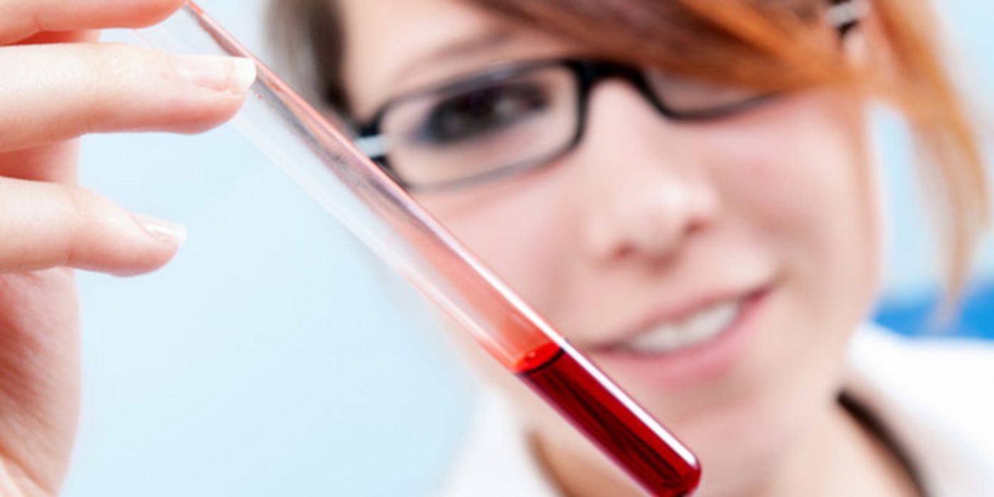 Großaufnahme Blutprobenröhrchen im Vordergrund, gehalten von Laborantin, rothaarig, dunkele Brille im Hintergrund (unscharf zu sehen), die lächelnd auf das Röhrchen schaut