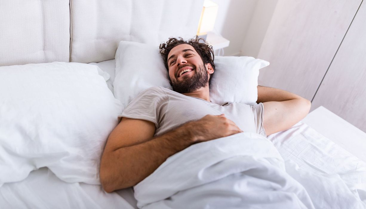 Junger Mann mit braunen Locken liegt im Bett und lächelt, ist ausgeschlafen.