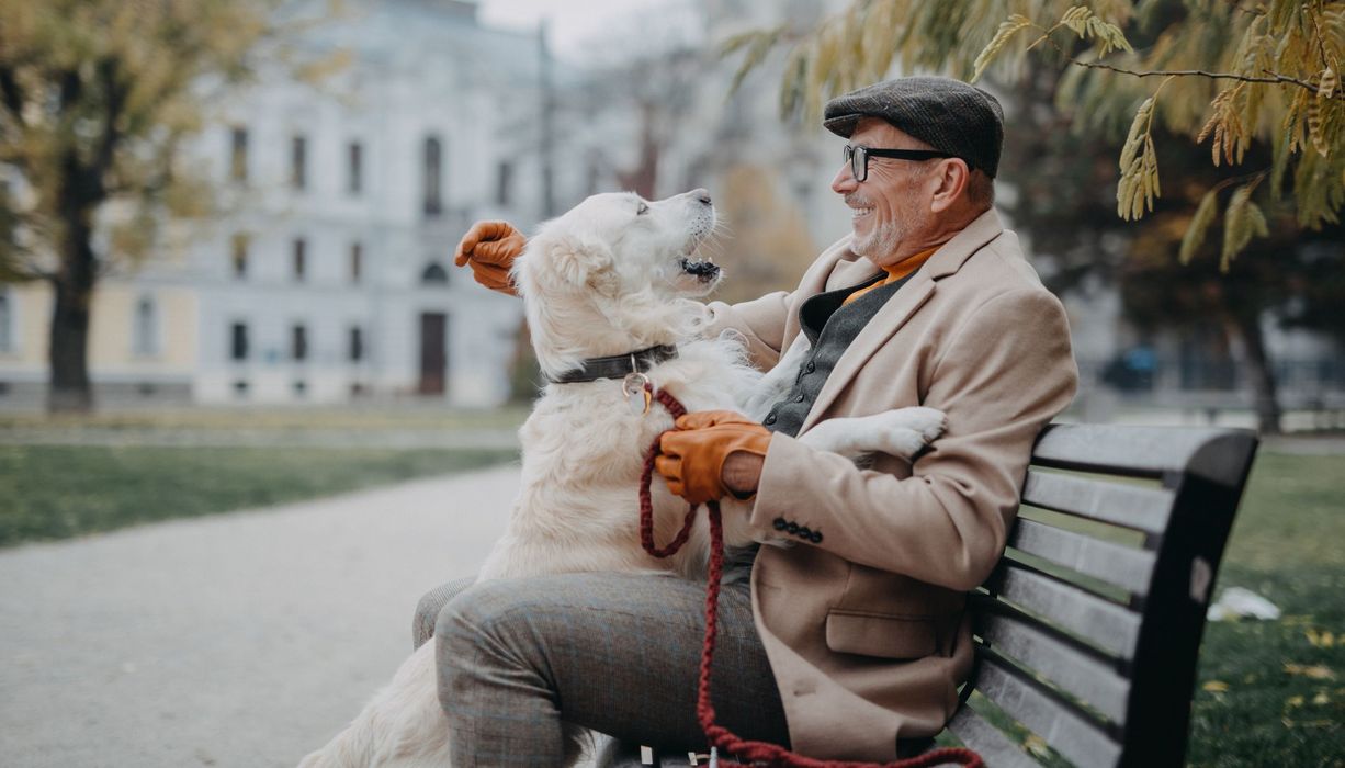 Älterer Mann, sitzt auf einer Bank, sein Hund stützt sich auf seinen Schoß.