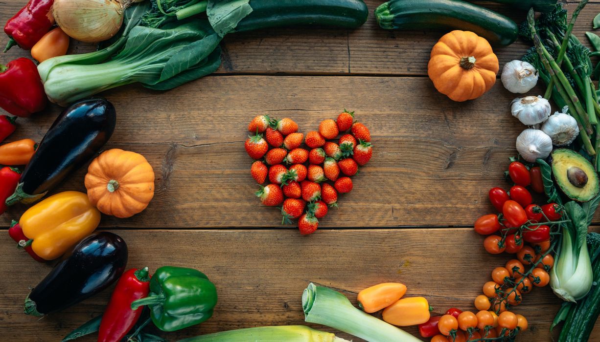 Auf einem Tisch liegen Erdbeeren als Herz angeordnet. Verschiedenes Gemüse bildet einen Kreis drumherum. 