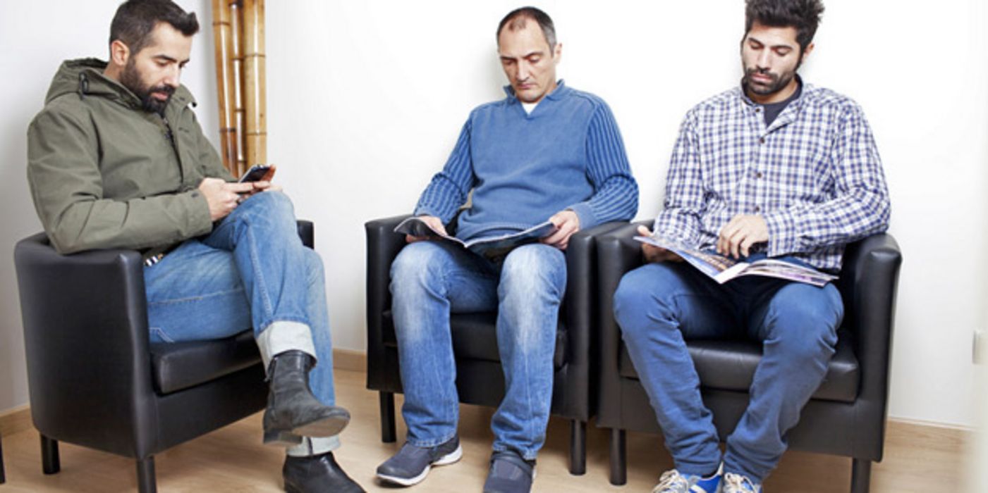 Drei junge Männer in einem Wartezimmer; einer spielt an seinem Handy, die anderen zwei lesen