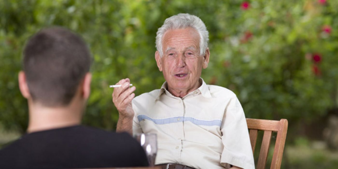 Älterer Mann draußen am Tisch rauchend, gegenüber mit dem Rücken zur Kamera