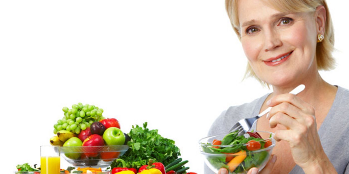 Blonde Frau um die 50 isst ein Schälchen mit gemischtem Salat; auf dem Tisch Obstschale und darum herum verschiedene Gemüse, eine Müslischale und ein Glas Orangensaft