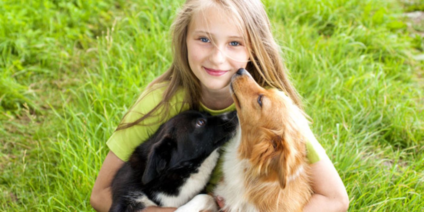 Junges Mädchen hockt im Gras und umarmt zwei mittelgroße Hunde