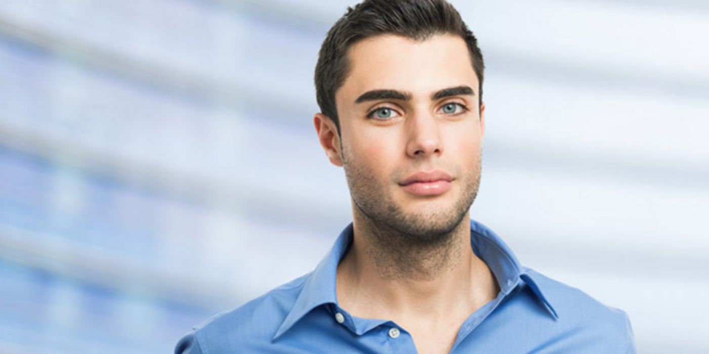 Melancholisch schauender jüngerer dunkelhaariger Mann, blaue Augen, hellblaues Hemd, im Hintergrund schemenhaft ein Bürohaus