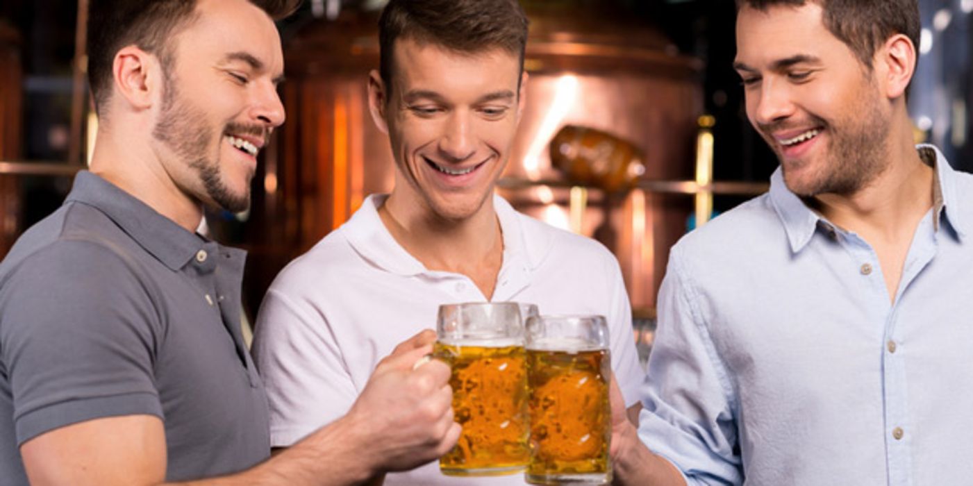 Drei Männer in den 20ern stoßen in einer Kneipe lachend mit Bierhumpen an