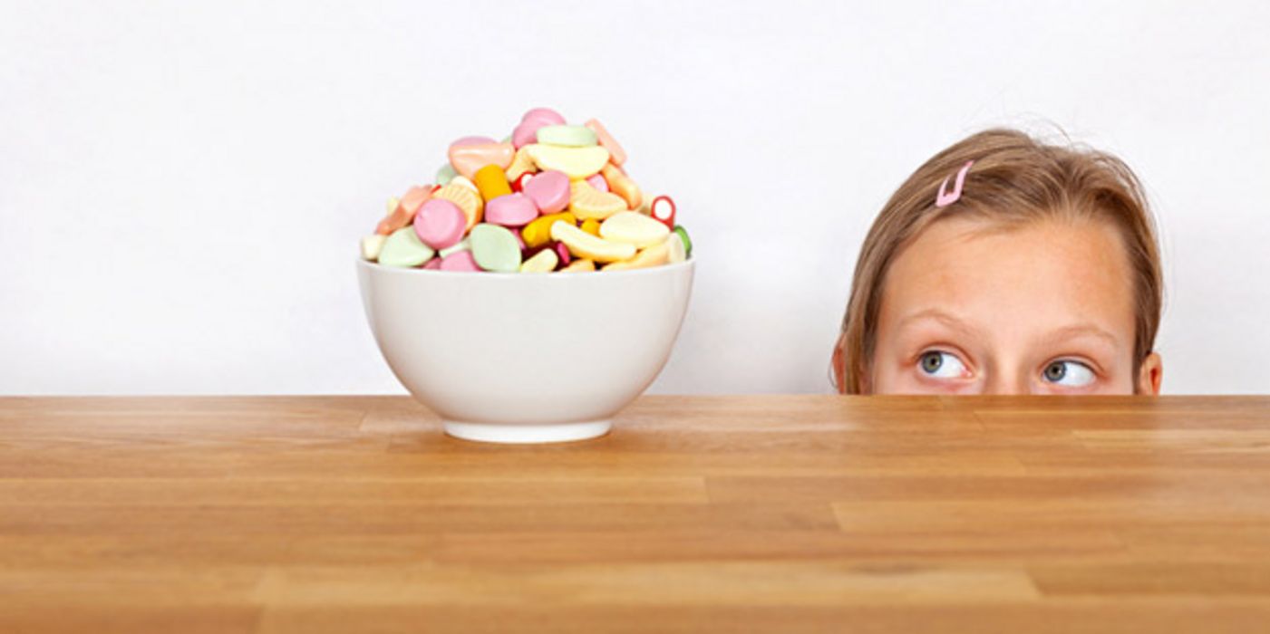 Tisch, darauf weißes Schälchen mit pastellfarbenen Süßigkeiten, Mädchen, nur bis Nasenansatz zu sehen, schaut mit verdrehten Augen hin
