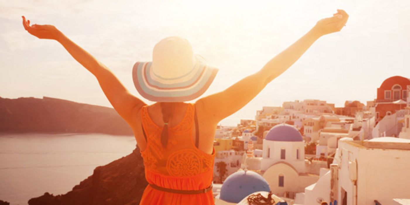 Junge Frau mit einem Sonnenhut, steht mit dem Rücken zu einer griechischen Stadt und breitet die Arme aus.