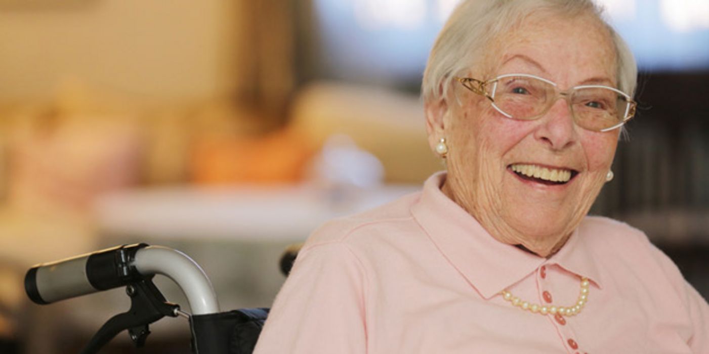 Lachende Seniorin im Rollstuhl sitzend, rosa Polopulli, graue, kurze Haare, Perlenohringe, -kett und schicke Brille 