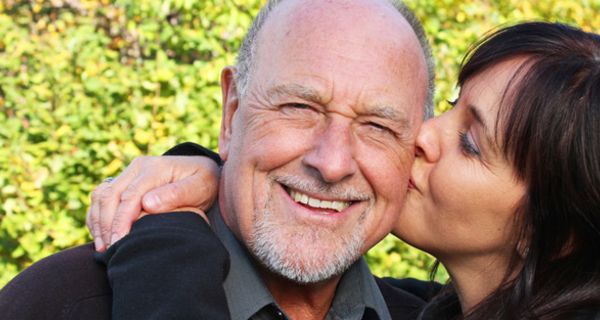 Portrait attraktives Paar Mitte 50; Mann mit grauem Dreitagebart lacht in die Kamera und wird von Frau auf die Wange geküsst
