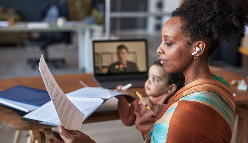 Eine junge Mutter hat ihr Kind auf dem Arm. Sie hält ein Blatt in der Hand und hat Köpfhörer auf. Gleichzeitig läuft im Hintergrund ein Videoanruf.