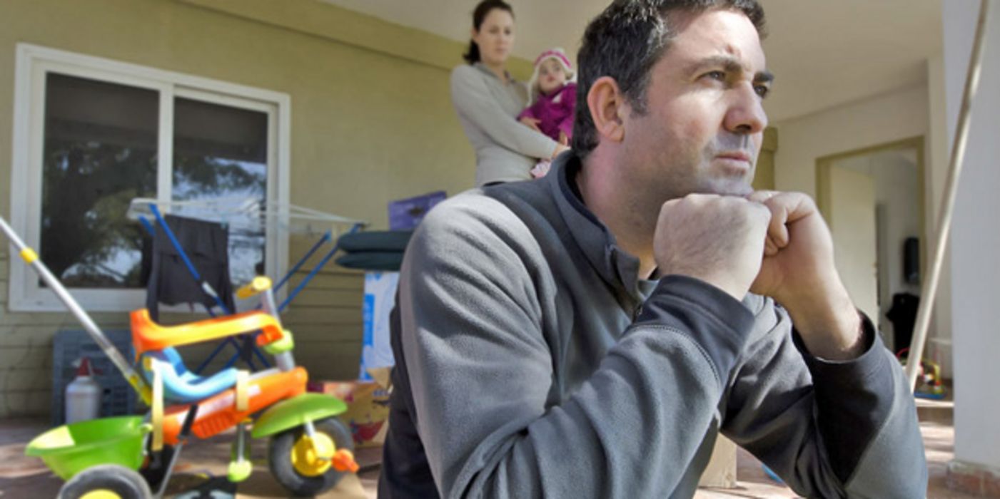 Resigniert-gestresster Mann im Vordergrund, im Hintergrund Frau mit Kleinkind auf dem Arm, unaufgeräumtes Zimmer