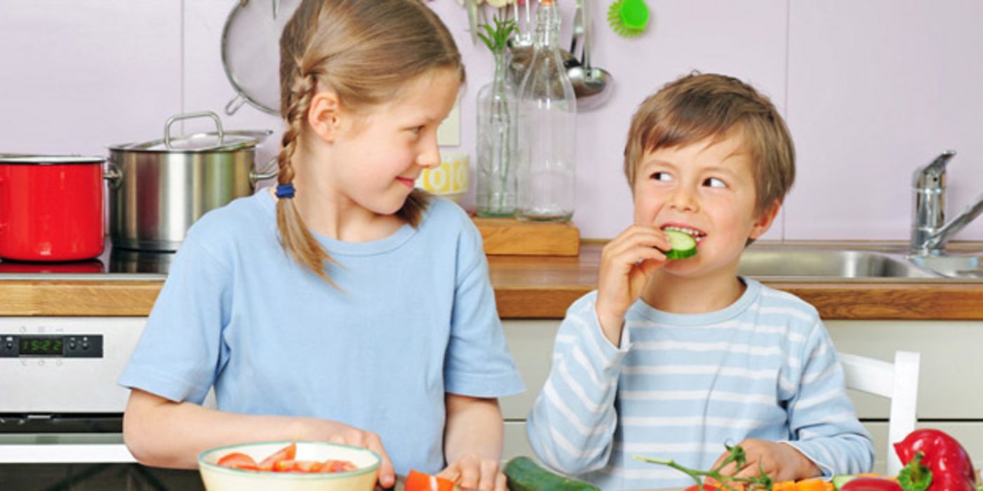 Geschwisterpaar beim Gemüseschneiden, Junge isst eine Gurkenscheibe und schaut seine ältere Schwester an