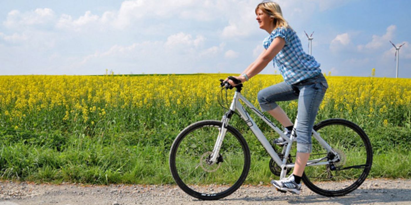 Junge Frau fährt mit dem Fahrrad einen Feldweg entlang; im Hintergrund ein Rapsfeld und Windräder