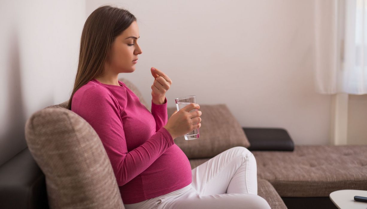 Schwangere Frau hält in der einen Hand ein Schmerzmittel und in der anderen Hand ein Glas Wasser.