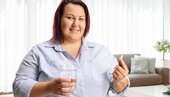 Eine Frau mit Übergewicht hält lächelnd eine Tablette und ein Glas Wasser in der Hand.