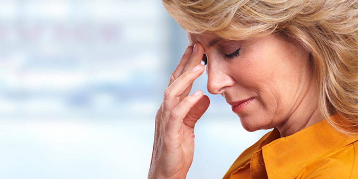 Rechter Bildrand Profil: Frau um die 60, blond, gelbe Bluse, Fingerspitzen der einen Hand fasst an schmerzende Stirn