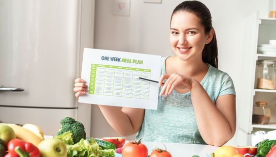 Eine junge Frau zeigt mit einem Stift auf einen Wochenspeiseplan. Vor ihr liegt Obst und Gemüse.